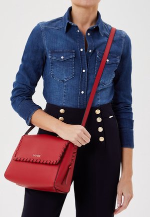 Červená kožená kabelka s designem šitých okrajů, zlatým logem a nastavitelným ramenním popruhem, držená osobou v džínové košili.