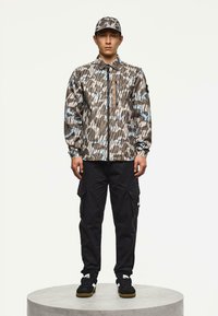 Uomo con giacca a fantasia marrone, beige e blu abbinata a un cappello, pantaloni cargo neri e sneakers nere, in piedi su una piattaforma rotonda.