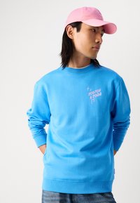 Lichtblauw katoenen sweatshirt met ribgebreide boorden en hals, met een roze grafisch logo op de borst. Gecombineerd met een roze pet.