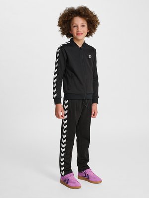 Kind mit lockigem Haar, das eine schwarze Reißverschlussjacke und Hose mit weißen Chevron-Streifen trägt, steht selbstbewusst in lila Turnschuhen vor einfachem Hintergrund.