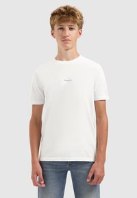 Tiener met lichtbruin haar, gekleed in een wit "BALLIN" t-shirt en blauwe jeans, staand tegen een vlakke grijze achtergrond.