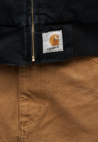 Κοντινό πλάνο φερμουάρ μαύρου μπουφάν Carhartt και λογότυπο πάνω από την τσέπη καφέ παντελονιού με εμφανείς ραφές.