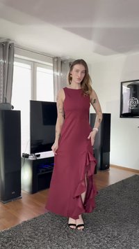 Dlouhé vínové šaty s přiléhavým živůtkem a rozparkem s volánky, vyrobené z hladké látky. Stylizované s černými sandály s otevřenou špičkou, zasazeno do moderního interiéru.