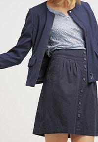 Meri zippered jakki navy berwarna dengan kain bertekstur dan leher bulat, dipadukan dengan rok A-line berciri pinggang tinggi yang sepadan, dilengkapi dengan kancing di bagian depan dan lipatan.