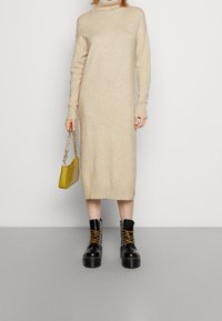 Béžové pletené šaty s rolákem a žebrovanými manžetami, bočními rozparky, v kombinaci s černými platformovými bojovými botami a malou olivově zelenou kabelkou.