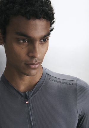 Giovane uomo con capelli corti e ricci che indossa una maglia da ciclismo Castelli grigia con zip, guarda leggermente verso il basso e di lato.