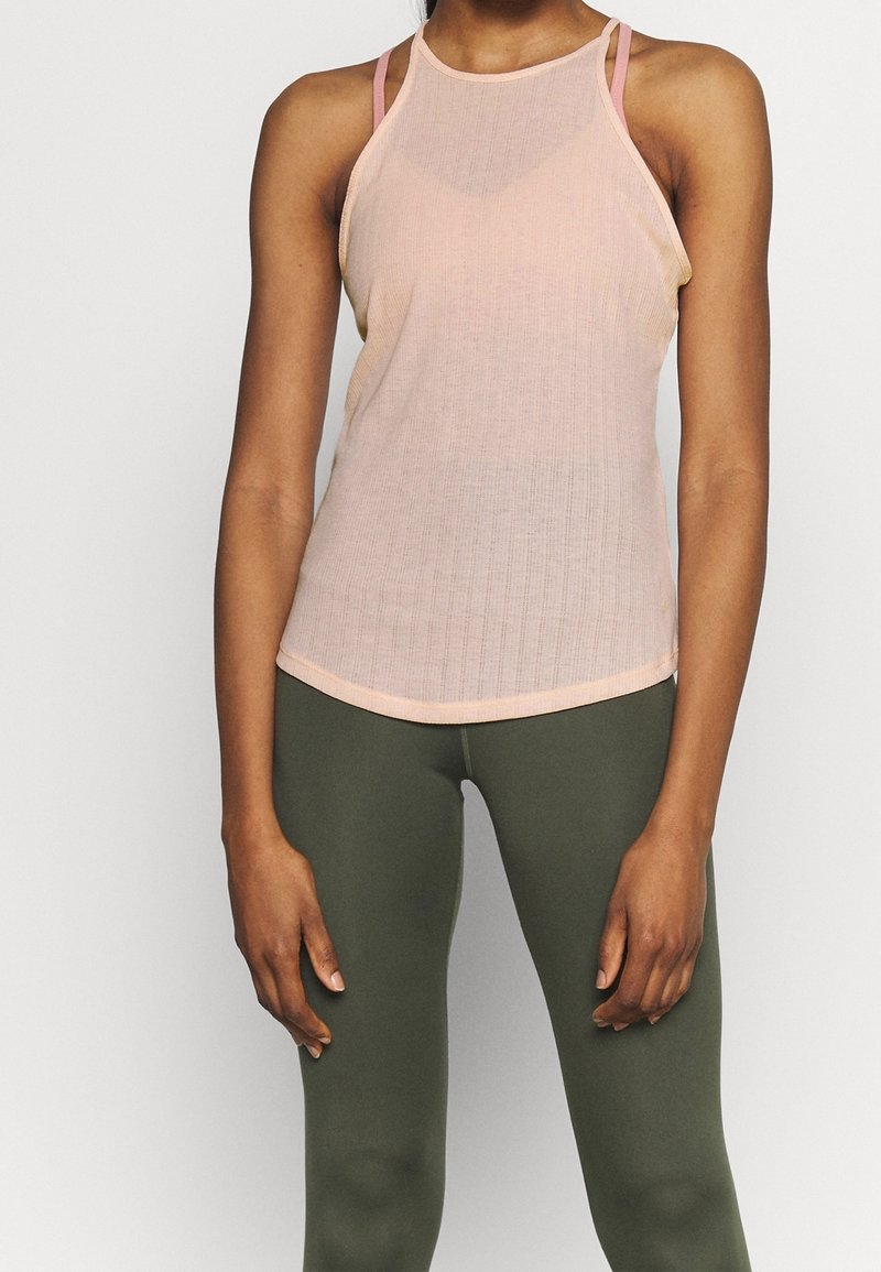 Femme portant un débardeur côtelé rose pâle sans manches et des leggings ajustés vert olive, debout devant un fond uni clair.