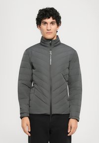 Ung mand med krøllet hår iført en mørk grå quiltet jakke med lynlås og EA7 Emporio Armani-logo på lynlåsen.