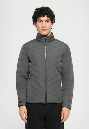 Young man with curly hair wearing a dark gray quilted zip-up jacket with EA7 Emporio Armani logo on the zipper.
