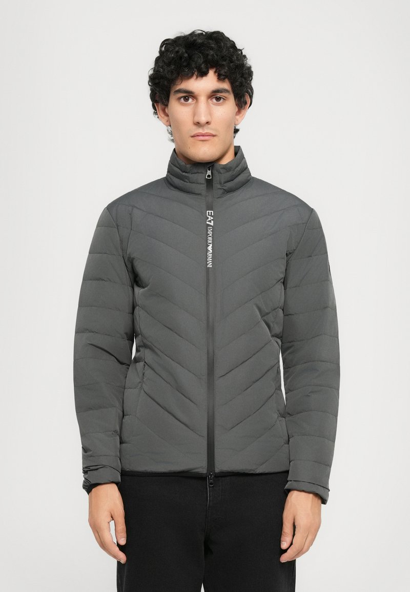 Ung mand med krøllet hår iført en mørk grå quiltet jakke med lynlås og EA7 Emporio Armani-logo på lynlåsen.