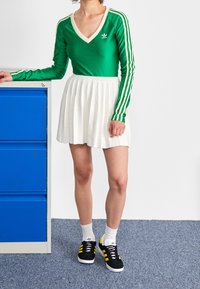 Camiseta verde de manga larga con ribete blanco y tres rayas laterales, combinada con una falda blanca plisada y zapatillas negras con detalles amarillos.