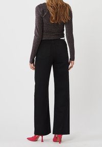 Frau in einem glitzernden Langarmtop, hochgeschnittenen, weiten schwarzen Hosen und rosa High Heels, die mit dem Rücken zu uns steht.