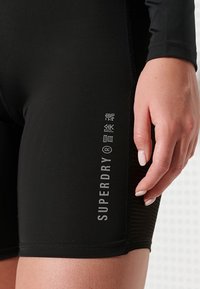 Svarta idrottsshorts med en åtsittande design, tillverkade av ett glatt material. Har en vertikal logotryck på vänster sida och texturdetaljer.
