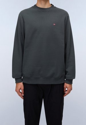 Man draagt een donkergrijze sweater met ronde hals en een kleine Noorse vlag geborduurd op de linkerborst, gecombineerd met een zwarte broek.