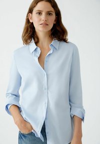 Light blue button-up shirt made of smooth fabric, featuring a collar, long sleeves rolled at the cuffs, and a relaxed fit.