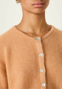 Pehmea virsikuvärvi villane kardigan ümmarguse kaelusega, viie pärlilaadse nööbiga ja pehme tekstuuriga. Kerge ka mugava lõikega.