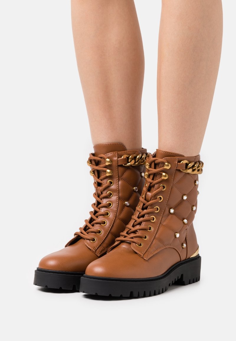 Bottes marron matelassées à lacets avec garnitures dorées, accents de perles, semelle noire épaisse et cheville rembourrée pour le soutien.