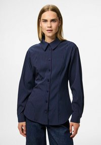 Woman wearing a navy blue button-up shirt with thin red vertical stripes, paired with dark blue jeans, standing against a plain background.