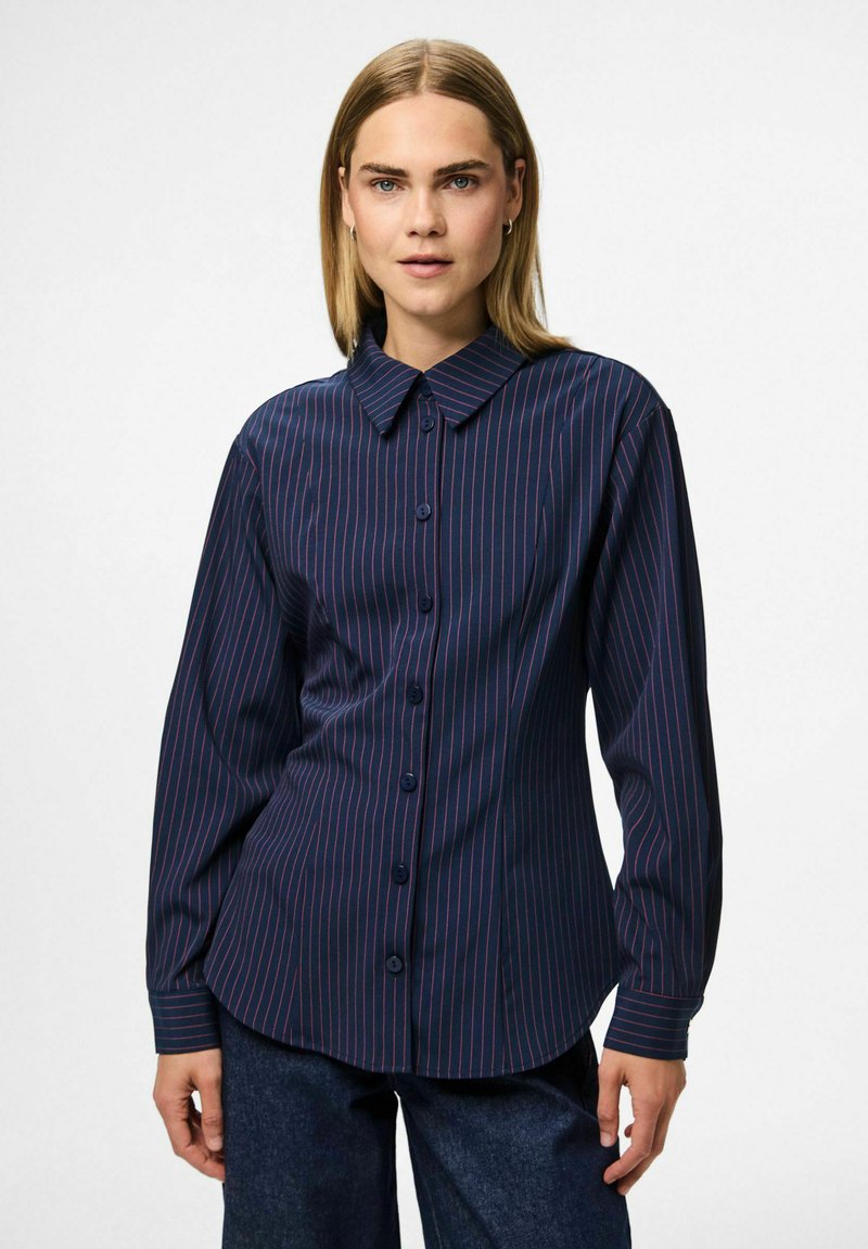 Woman wearing a navy blue button-up shirt with thin red vertical stripes, paired with dark blue jeans, standing against a plain background.