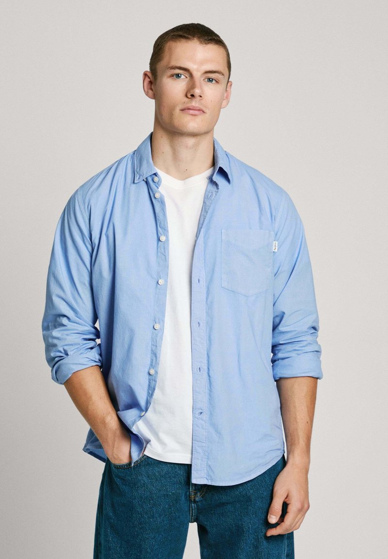 Ljusblå skjorta med knappar, med ett bröstfick, bärs över en vit T-shirt. Modellen matchar den med denimjeans. Slät tyg, avslappnad passform.