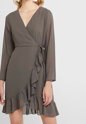 Vrouw draagt een grijze, lange mouw, wikkeljurk met geplooide zoom en zijdetail van een strik, tegen een effen achtergrond.