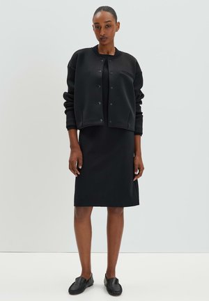 Schwarze Sweatshirtjacke mit Druckknöpfen und entspannter Passform, über einem figurbetonten schwarzen Kleid getragen. Schwarze Slipper runden das Outfit ab.