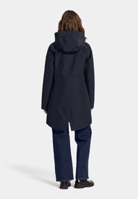 Marinfärgad vattentät parka med stor huva, rak skärning och detaljer med ventiler bak; kombinerat med löst sittande mörk denimbyxor och svarta skor.