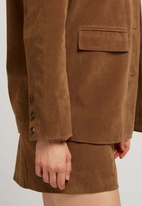 Blazer en daim marron avec un col à revers, une poche poitrine simple et des poignets à trois boutons, associé à une jupe courte coordonnée.