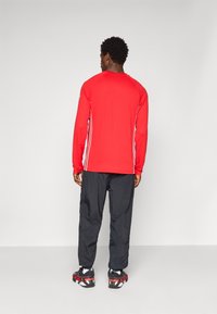 Red long-sleeve top with white side stripes, paired with black, loose-fitting pants and black-red athletic shoes, viewed from the back.