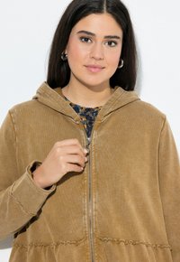 Felpa con zip oversize in bronzo con una texture morbida, dotata di cappuccio, orlo strappato e polsini a coste. Il modello sta tenendo la zip.