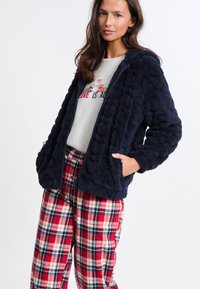 Námořnická modrá fleecová bunda s texturou, kapucí, zipovým uzávěrem a dvěma bočními kapsami, spárovaná s červenými kostkovanými pyžamovými kalhotami.