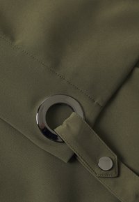 Tessuto verde oliva con una superficie liscia, caratterizzato da un anello grigio metallico e un dettaglio di chiusura a pressione. Cucitura visibile lungo il bordo.