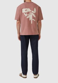 Kurzes, rosa Baumwoll-T-Shirt mit einem großen weißen Blumenmuster auf dem Rücken; kombiniert mit dunklen Denim-Hosen und lässigem Schuhwerk.