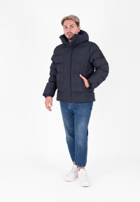 Zwarte pufferjack met een hoge kraag en capuchon, voorzien van een ritssluiting, zijzakken en een logo op de mouw, gedragen met blauwe spijkerbroeken.