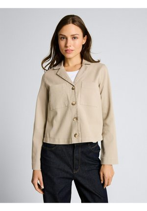 Frau trägt eine beige Jacke mit Knöpfen und zwei Brusttaschen über einem weißen Oberteil und dunkelblauen Jeans, steht vor einem hellgrauen Hintergrund.