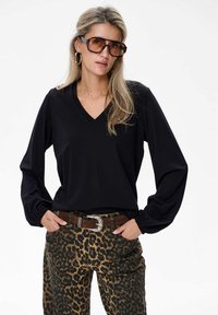 Schwarzes Langarmoberteil mit V-Ausschnitt, kombiniert mit Hosen im Leopardenmuster. Das Outfit umfasst einen braunen Gürtel mit einer silbernen Schnalle und übergroße Sonnenbrillen.