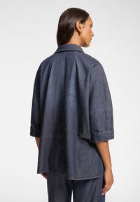 Camicia in denim con vestibilità rilassata, colore blu scuro, maniche corte arrotolate e dettaglio con piega sul retro, che mette in mostra una texture morbida e cuciture sottili.