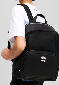 Black textured backpack with a zipped pocket featuring a cartoon face logo. Shoulder straps and top handle for carrying options.