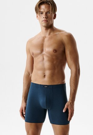 Młody mężczyzna o krótkich blond włosach, ubrany w ciemnoniebieskie bokserki, stoi na gładkim jasnym tle.