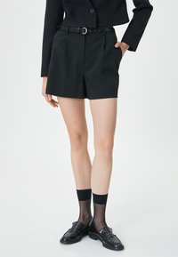 Pantaloni corti neri su misura con una texture liscia, dotati di una cintura ampia, passanti per la cintura e una cintura con anello in metallo. Abbinati a mocassini neri e calze.
