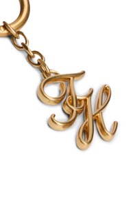 Μπρελόκ από μέταλλο σε χρυσό τόνο με διαπλεκόμενα καλλιγραφικά γράμματα "T" και "H", προσδεμένα με διακοσμητικό κρίκο αλυσίδας.
