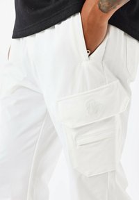 Pantalones de carga blancos con una textura en la tela, que cuentan con grandes bolsillos de parche y un sutil bordado decorativo. Diseñados con un ajuste relajado.