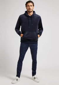 Sudadera de forro polar azul marino con un bolsillo tipo canguro, capucha con cordón y puños acanalados, combinada con jeans oscuros ajustados y zapatillas blancas.