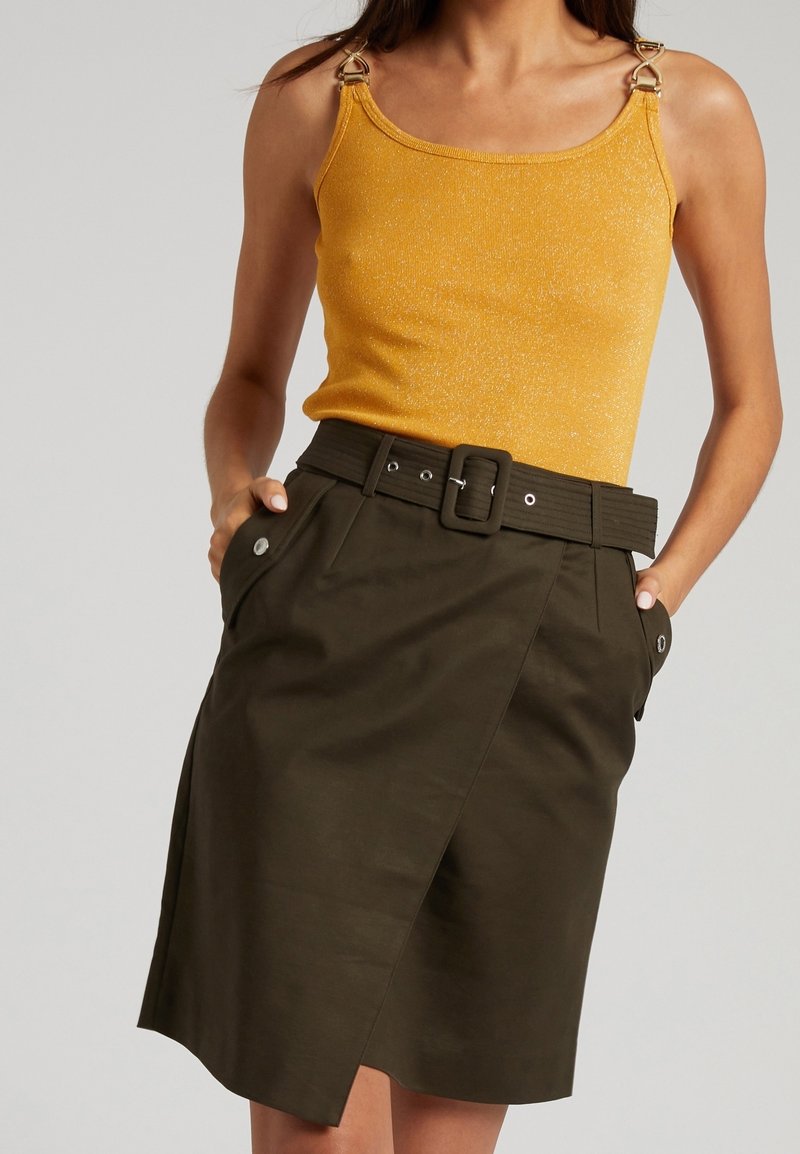 Femme portant un haut jaune sans manches avec des détails anneau doré et une jupe portefeuille taille haute couleur olive foncé avec une large ceinture et des poches.