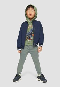 Marine bomberjack met gestreepte manchetten, groene hoodie met dinosaurustoon, lichtgroene broek en zwarte sneakers met neonaccenten.