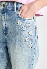 Lichtblauwe jeans van denim met bloemembroiderie op de dij, voorzien van vijf witte madeliefachtige bloemen. Klassiek model met vijf zakken, hoge taille.
