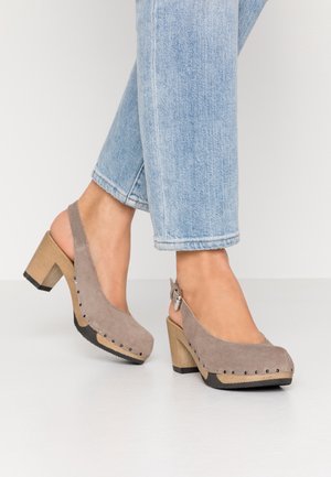 Jasnoniebieskie dżinsy oraz taupe zamszowe buty na słupku typu slingback z drewnianą podeszwą i zdobione ćwiekami, noszone na białej podłodze.