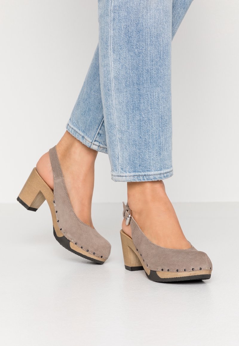 Jean bleu clair et chaussures slingback en daim taupe à talon bloc avec semelle en bois et bordure cloutée portés sur un sol blanc.