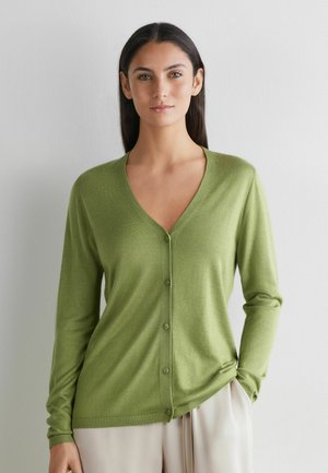 Kobieta o długich ciemnych włosach, ubrana w jasnobrązowy kardigan zapinany na guziki i beżowe spodnie, stoi na tle jednolitego białego tła.