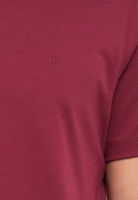 Burgundi színű pamut póló rövid ujjakkal és kerek nyakkal. Diszkrét hímzett "MK" logó található a mellkason. Puha tapintású.
